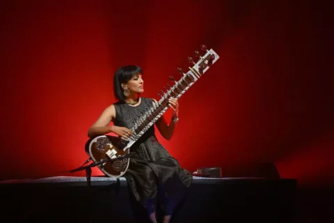 Hráčka na sitar sedí na pódiu, osvetlená teplým červeným pozadím. Má na sebe trblietavé čierne šaty a veľké náušnice, ktoré vytvárajú pokojnú atmosféru.