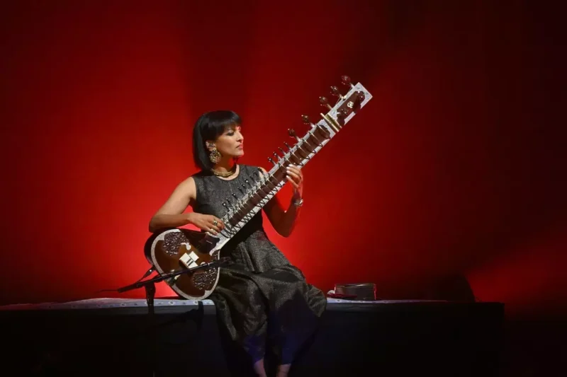 Hráčka na sitar sedí na pódiu, osvetlená teplým červeným pozadím. Má na sebe trblietavé čierne šaty a veľké náušnice, ktoré vytvárajú pokojnú atmosféru.