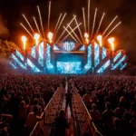 Dav na nočnom hudobnom festivale, žiarivé svetlá pódia a ohňostroj osvetľujú oblohu. Energická atmosféra s veľkou video projekciou.