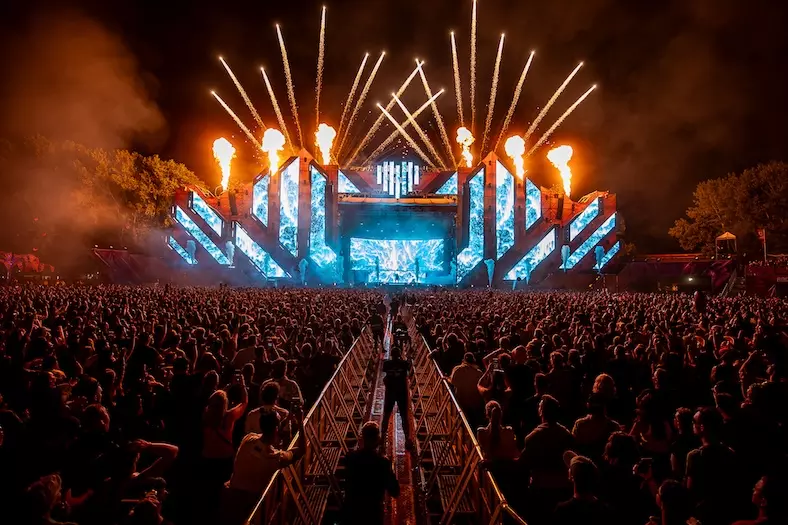 Dav na nočnom hudobnom festivale, žiarivé svetlá pódia a ohňostroj osvetľujú oblohu. Energická atmosféra s veľkou video projekciou.