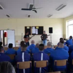 Hudobníci s gitarami hrajú v miestnosti s niekoľkými sediacimi ľuďmi v modrých uniformách. Prostredie je neformálne, čo podporuje uvoľnenú a pozornú atmosféru.