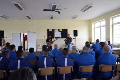 Hudobníci s gitarami hrajú v miestnosti s niekoľkými sediacimi ľuďmi v modrých uniformách. Prostredie je neformálne, čo podporuje uvoľnenú a pozornú atmosféru.