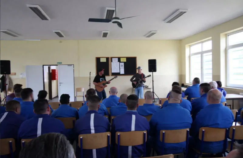 Hudobníci s gitarami hrajú v miestnosti s niekoľkými sediacimi ľuďmi v modrých uniformách. Prostredie je neformálne, čo podporuje uvoľnenú a pozornú atmosféru.