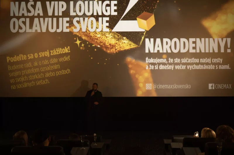 V slabo osvetlenom kine stojí človek a prihovára sa publiku. Na veľkej obrazovke sa v zlatej grafike zobrazuje oslavný odkaz k druhému výročiu VIP salónu, čím vytvára slávnostnú atmosféru.