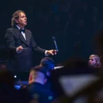 Dirigent v smokingu vedie orchester na slabo osvetlenom pódiu a vytvára atmosféru sústredenia a očakávania. Neďaleko je vidieť huslistu.