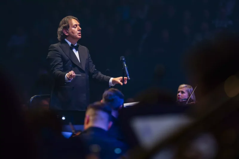 Dirigent v smokingu vedie orchester na slabo osvetlenom pódiu a vytvára atmosféru sústredenia a očakávania. Neďaleko je vidieť huslistu.