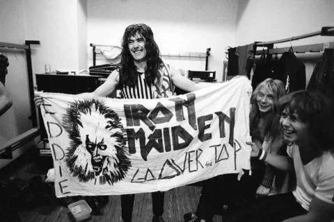 Čiernobiela fotografia usmievavého muža, ktorý drží transparent s nápisom „Iron Maiden“ a zobrazuje monštruóznu tvár. Neďaleko sedia ďalší dvaja a všetci vyzerajú šťastne a energicky v zákulisí.