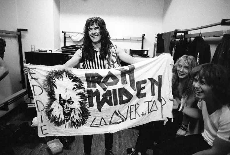 Čiernobiela fotografia usmievavého muža, ktorý drží transparent s nápisom „Iron Maiden“ a zobrazuje monštruóznu tvár. Neďaleko sedia ďalší dvaja a všetci vyzerajú šťastne a energicky v zákulisí.