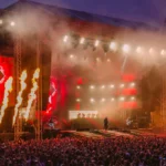 Veľký vonkajší koncert za súmraku s davom. Z pódia šľahajú jasne oranžové plamene obklopené červenými svetlami a veľkými videoobrazovkami, ktoré vytvárajú elektrizujúcu atmosféru.
