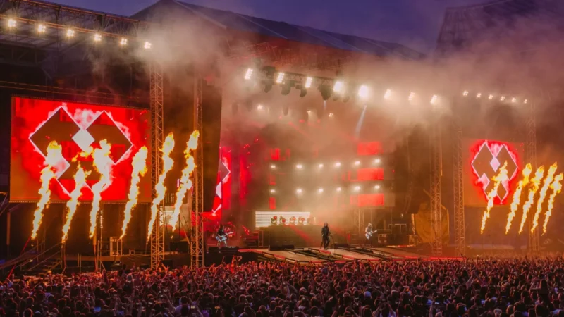 Veľký vonkajší koncert za súmraku s davom. Z pódia šľahajú jasne oranžové plamene obklopené červenými svetlami a veľkými videoobrazovkami, ktoré vytvárajú elektrizujúcu atmosféru.