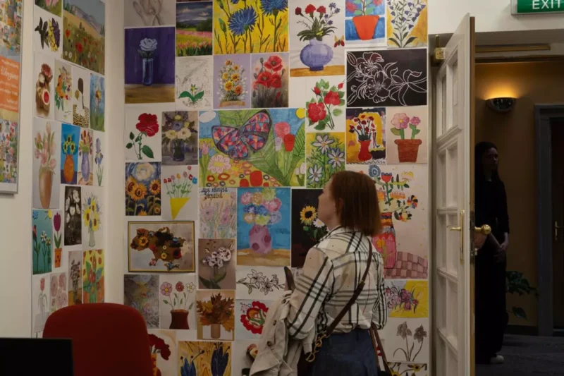 Žena s ryšavými vlasmi a v kockovanej košeli obdivuje farebnú stenu plnú rôznych kvetinových obrazov a náčrtov, ktoré vytvárajú živú a umeleckú expozíciu.