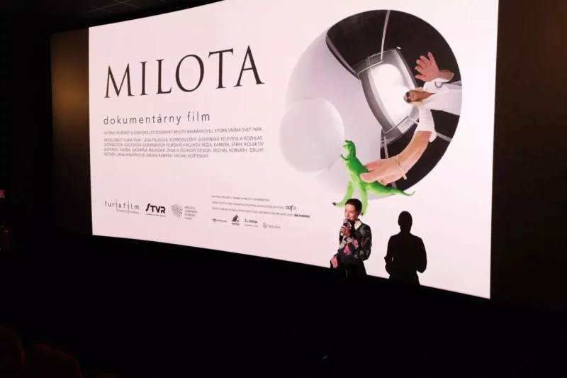 Muž stojí na pódiu kina a hovorí do mikrofónu pred veľkou obrazovkou, na ktorej je zobrazený plagát k dokumentárnemu filmu „Milota“.