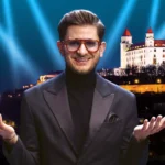 Muž v obleku s okuliarmi stojí s úsmevom a otvorenou náručou na pozadí osvetleného hradu v noci pod oblohou plnou jasných reflektorov.