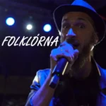 Bradatý muž v klobúku vášnivo spieva do mikrofónu pod modrými svetlami pódia. Naľavo je výrazne zobrazený text „FOLKLÓRNA“.