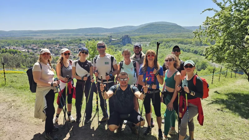 Skupina turistov šťastne pózuje na slnečnom chodníku s trekingovými palicami v rukách. Sú obklopení sviežimi zelenými kopcami a rozľahlou vidieckou krajinou.