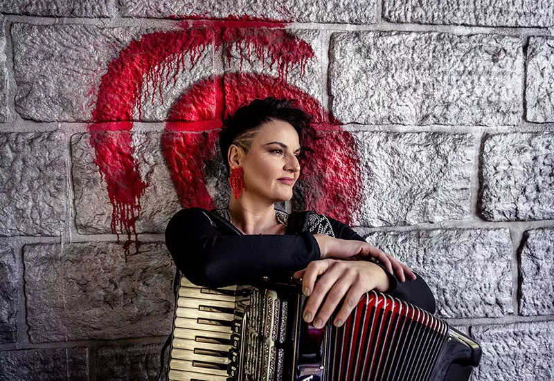 Osoba s krátkymi tmavými vlasmi sa opiera o textúrovanú kamennú stenu a drží v ruke harmoniku. Na stene je červený vzor, ​​ktorý jej dodáva umelecký nádych. Výraz pôsobí zamyslene.