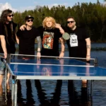 Štyria muži stoja vo vode okolo plávajúceho pingpongového stola a usmievajú sa. Sú ležérne oblečení, v pozadí svieža zeleň a jazero.