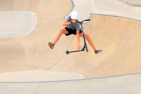 Mladý človek v bielej prilbe predvádza vzdušný trik na kolobežke s natiahnutými nohami v betónovom skateparku. Obrázok zobrazuje dynamickú akciu.