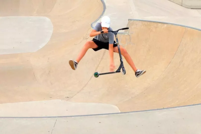 Mladý človek v bielej prilbe predvádza vzdušný trik na kolobežke s natiahnutými nohami v betónovom skateparku. Obrázok zobrazuje dynamickú akciu.