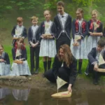 Skupina detí a muž, oblečení vo viktoriánskom oblečení, spúšťajú drevené hračkárske loďky do hmlistého lesného potoka, čo vyvoláva pocit nostalgie a radosti.