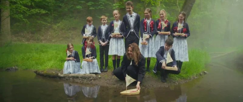 Skupina detí a muž, oblečení vo viktoriánskom oblečení, spúšťajú drevené hračkárske loďky do hmlistého lesného potoka, čo vyvoláva pocit nostalgie a radosti.