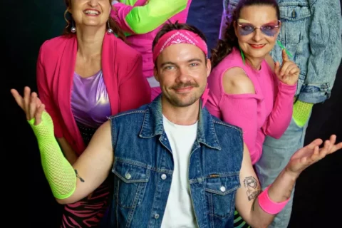 Skupina piatich dospelých pózuje v žiarivých retro outfitoch z 80. rokov s neónovými farbami a džínsovinami a energicky sa usmieva na tmavom pozadí.