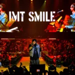 Umelec energicky spieva na pódiu obklopenom žiarivými svetlami. Na veľkej obrazovke sa zobrazuje nápis „IMT SMILE“ s dynamickými farbami a pod ňou jasajúci dav.