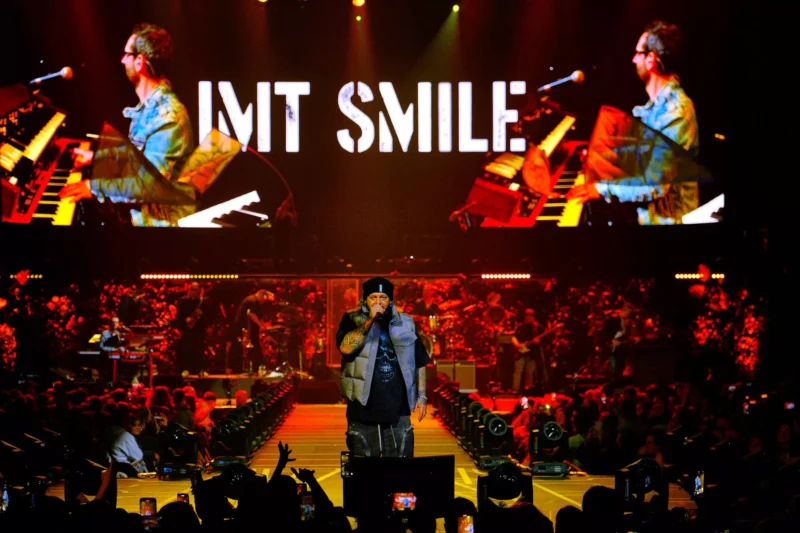 Umelec energicky spieva na pódiu obklopenom žiarivými svetlami. Na veľkej obrazovke sa zobrazuje nápis „IMT SMILE“ s dynamickými farbami a pod ňou jasajúci dav.