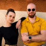 Žena v čiernom tope sa ležérne opiera o muža s bradou, ktorý má na sebe slnečné okuliare a žlté tričko, obaja sa usmievajú na pozadí dvojfarebnej textúrovanej steny.