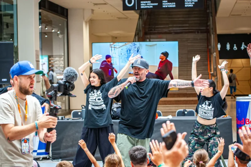 Živá scéna v nákupnom centre s účinkujúcimi na pódiu, ktorí zaujmú dav. Muž s mikrofónom a dve ženy vedú nadšený tanec so zdvihnutými rukami.