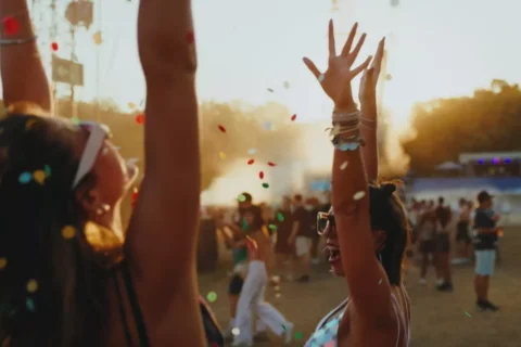 Ľudia oslavujú na vonkajšom hudobnom festivale so zdvihnutými rukami a lietajúcimi konfetami. V pozadí zapadá slnko a vytvára vrúcnu a radostnú atmosféru.