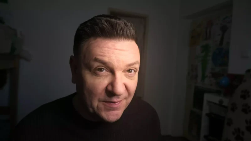Muž v tmavej miestnosti sa s miernym úsmevom pozerá do kamery. Má krátke tmavé vlasy a oblečenú čiernu košeľu. Pozadie je slabo osvetlené a na stene visia plagáty.