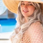 Usmievavá žena s dlhými blond vlasmi, ktorá má na sebe slamený klobúk so širokým okrajom a sieťovaný župan, stojí pri bazéne. Scéna je svetlá a letná.