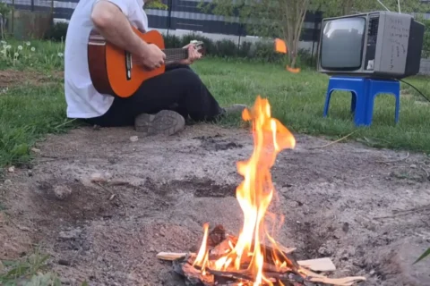 Osoba hrajúca na gitare sedí pri malom ohnisku na trávnatom pozemku, v pozadí je starý televízor na modrej stoličke, čo evokuje rustikálnu, uvoľnenú atmosféru.