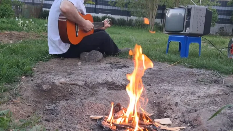Osoba hrajúca na gitare sedí pri malom ohnisku na trávnatom pozemku, v pozadí je starý televízor na modrej stoličke, čo evokuje rustikálnu, uvoľnenú atmosféru.