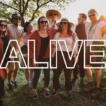 Skupina radostných ľudí v ležérnom oblečení si užíva slnečný deň vonku, obklopení zeleňou. Cez obrázok je nápis „ALIVE“, ktorý vyjadruje energiu a šťastie.