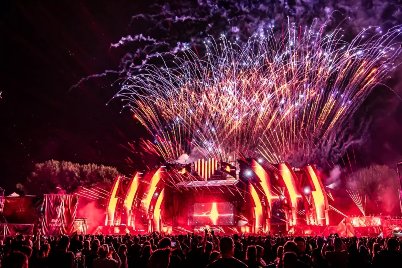 Živé pódium hudobného festivalu osvetlené červenými svetlami s ohňostrojmi explodujúcimi nad hlavou, ktoré zachytávajú živú a slávnostnú atmosféru. Siluety veľkého davu.