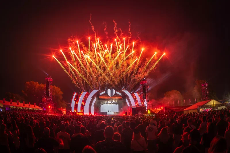 Živý hudobný festival v noci s veľkým davom stojacim pred osvetleným pódiom. Nad pódiom vybuchujú červené ohňostroje, ktoré vytvárajú energickú a slávnostnú atmosféru.