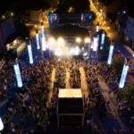 Letecký pohľad na živý nočný koncert s veľkým davom pred jasne osvetleným pódiom. Vysoké osvetlené stĺpy obklopujú námestie a vytvárajú slávnostnú atmosféru.