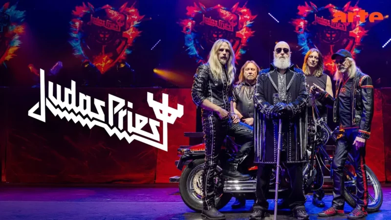 Heavymetalová kapela sebavedomo pózuje na pódiu s jasnými, farebnými svetlami. Členovia majú na sebe tmavé oblečenie s cvočkami. Je na ňom zobrazené logo „Judas Priest“. Dramatický a energický tón.
