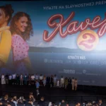 Na veľkom filmovom plátne je plagát k filmu „Kavej 2“ s dvoma usmievavými ženami, jednou v žltom, druhou v ružových bodkách. Pod ním stojí na javisku rad ľudí tvárou v tvár publiku v divadelnom prostredí. Atmosféra je slávnostná, pripomínajúca premiéru filmu.