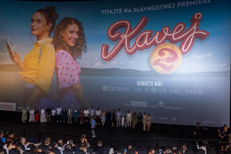 Na veľkom filmovom plátne je plagát k filmu „Kavej 2“ s dvoma usmievavými ženami, jednou v žltom, druhou v ružových bodkách. Pod ním stojí na javisku rad ľudí tvárou v tvár publiku v divadelnom prostredí. Atmosféra je slávnostná, pripomínajúca premiéru filmu.