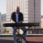 Človek hrá na klávesoch na streche, obklopený mestskými budovami pod jasnou oblohou. Scéna vyvoláva uvoľnenú a kreatívnu atmosféru.