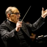 Starší dirigent s okuliarmi vedie orchester s výraznými gestami. Drží taktovku a je oblečený v čiernom obleku na tmavom pozadí.
