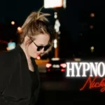 Žena so slnečnými okuliarmi a čiernou bundou stojí na pozadí nočného mesta. Nápis „Hypnotized“ od Nicky Mack vytvára štýlovú mestskú atmosféru.