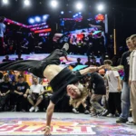 Breakdancer predvádza stojku na jednej ruke na živom pódiu, obklopený publikom a porotcami. Scéna je dynamická a energická.