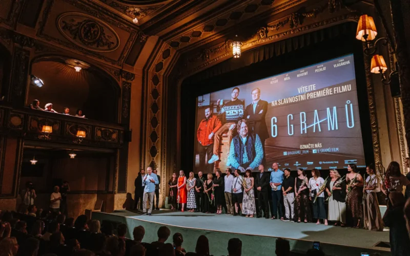 Rôznorodá skupina stojí na javisku v zdobenom kine pod plátnom premiéry filmu „6 gramov“, zatiaľ čo sediaci hostia sledujú premietanie v teplom, slávnostnom svetle.