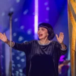 Speváčka v čiernom zamatovom oblečení dvíha obe ruky a usmieva sa na žiarivo modrofialovom pódiu, čím počas vystúpenia vyjadruje radosť a energiu.