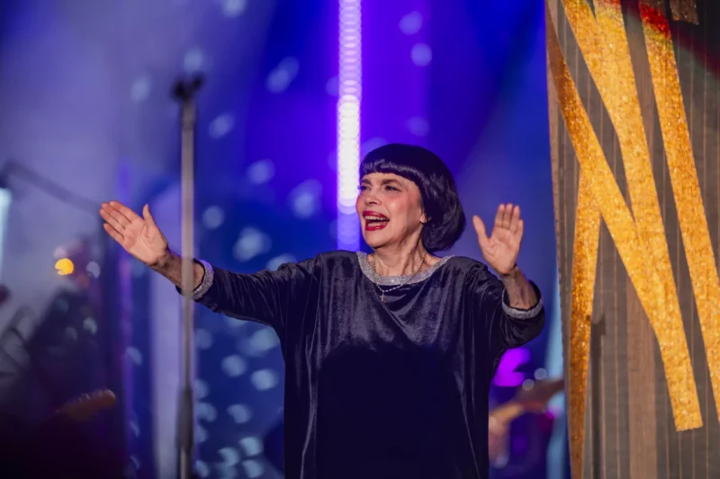 Speváčka v čiernom zamatovom oblečení dvíha obe ruky a usmieva sa na žiarivo modrofialovom pódiu, čím počas vystúpenia vyjadruje radosť a energiu.