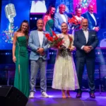 Osem účinkujúcich a moderátorov pózuje na jasne osvetlenom pódiu a usmieva sa s kvetmi pod farebnými obrazovkami na živom hudobnom festivale.